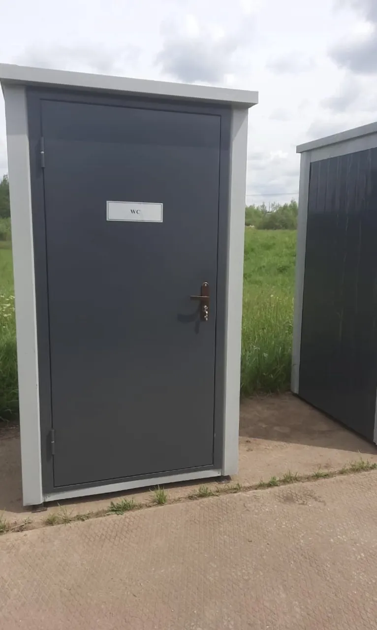 Серая туалетная кабина с металлической дверью и замком на бетонной площадке
